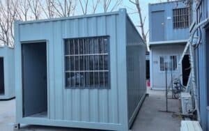 A light - blue container for refugee camp use, featuring a door and a window with metal bars, situated among other container structures.