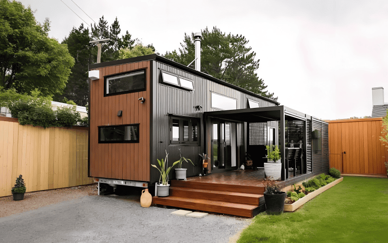 A high cube container home with a full mezzanine level. The home has a dark and wooden exterior, multiple windows, and a covered porch area with potted plants, situated in a fenced yard with greenery.