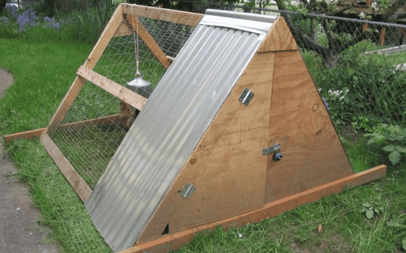 A triangular - shaped wooden pig house with a metal roof and wire mesh sides, placed on a grassy area.