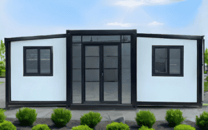 A modern modular container house with a black frame, large glass sliding door, and side windows, surrounded by small green shrubs under a clear blue sky.