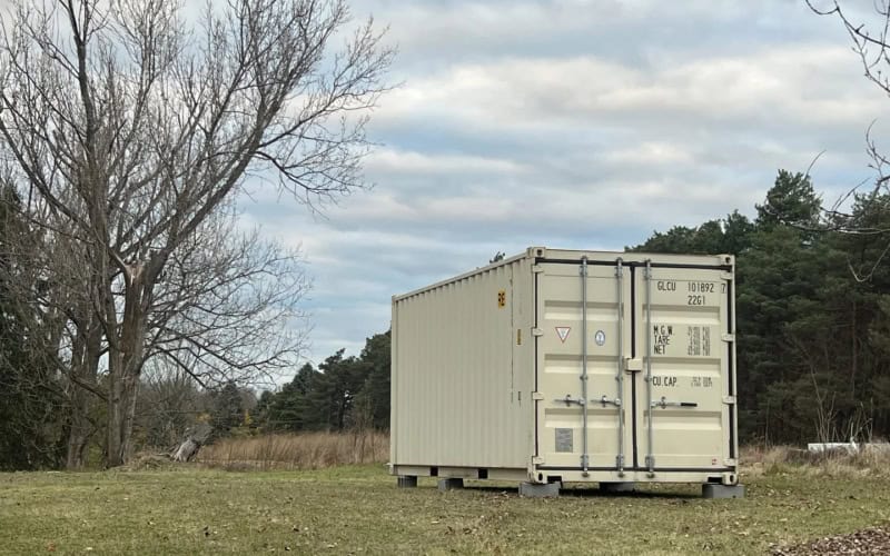 A beige shipping container set on a grassy area with trees in the background under a partly cloudy sky. The container has markings like "OCLI 101892 7 22G1" on its doors, indicating it's used for storage or other purposes.
