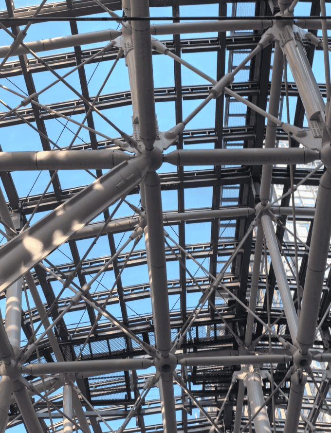 Interior view of a steel truss system showcasing complex structural design with intersecting beams and a clear blue sky visible through the roof.