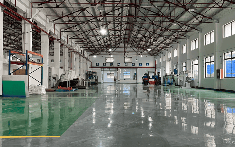 Wide-angle view of an industrial workshop interior with polished floors, red steel roof trusses, storage racks, and equipment, illuminated by overhead lights.