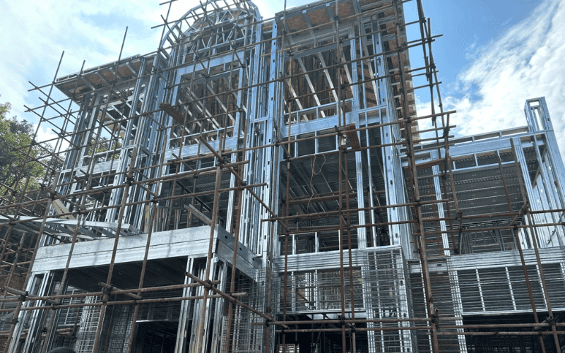 A steel-frame building under construction, surrounded by wooden scaffolding, showing intricate framework details under a partly cloudy sky.