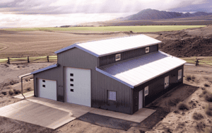 A steel barn with a modern design, featuring multiple garage doors, a sloped roof, and located in a rural setting with open fields and mountains in the background."