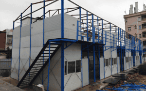 A prefabricated two-story modular structure under construction, featuring blue steel framing, external staircases, and partially assembled wall panels.