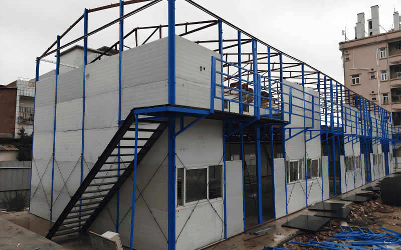 A prefabricated two-story modular structure under construction, featuring blue steel framing, external staircases, and partially assembled wall panels.