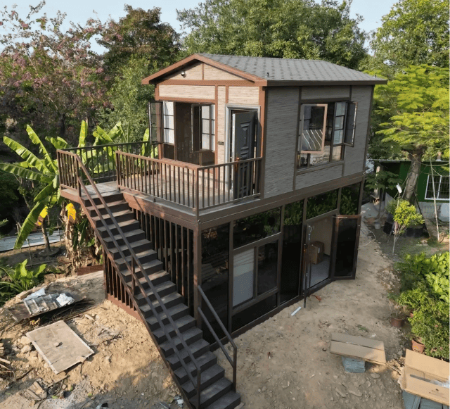 A modern prefabricated house with a balcony and staircase, built with modular construction, surrounded by greenery and construction materials.