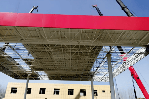 A steel canopy structure being installed with cranes, featuring a bright red top and a clear blue sky.