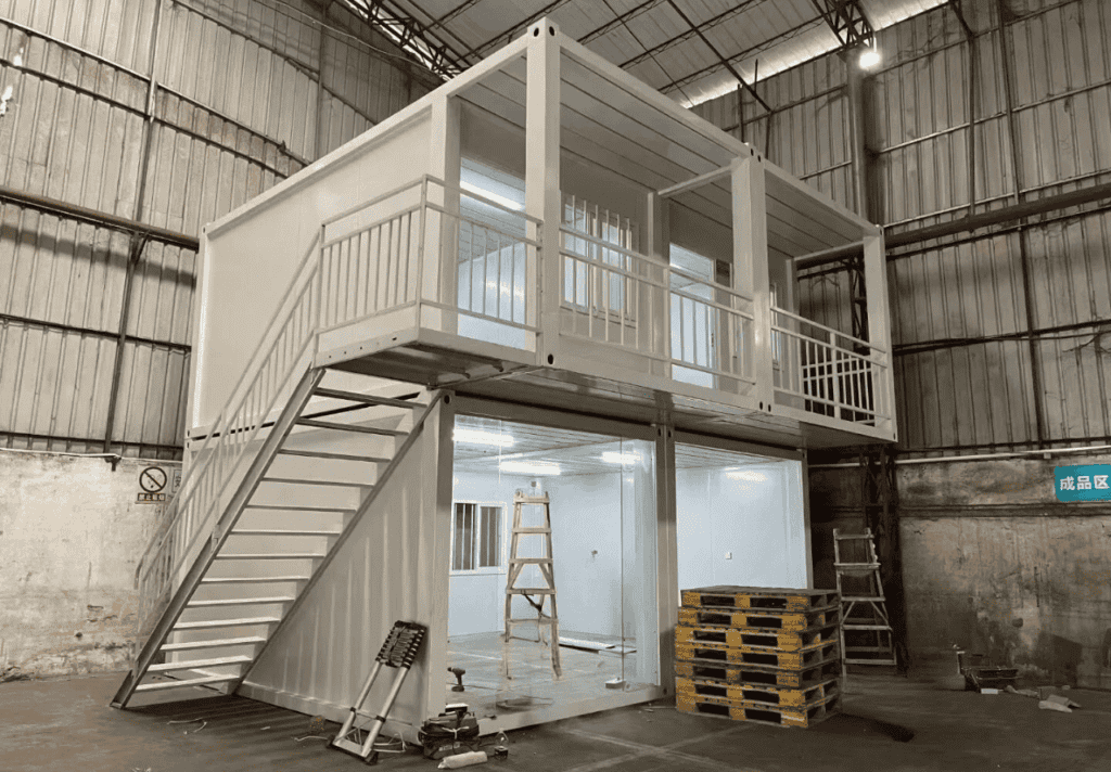 A two-story white container house with a staircase, balcony, and large glass windows, constructed inside an industrial warehouse.