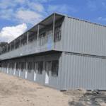 A two-story grey container housing complex featuring multiple units with separate doors, positioned on a construction site under clear skies.