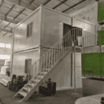 A compact two-story container house with a staircase and minimalist design, displayed inside a warehouse with surrounding equipment.