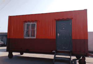 A red container house on wheels with a blue door and barred window, showcasing a portable and secure design.