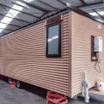 A beige container house on wheels with corrugated walls and a window, displayed indoors with a focus on mobility and functionality.