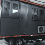 A dark gray container house on wheels with multiple windows and an air-conditioning unit, highlighting a compact and mobile living solution.