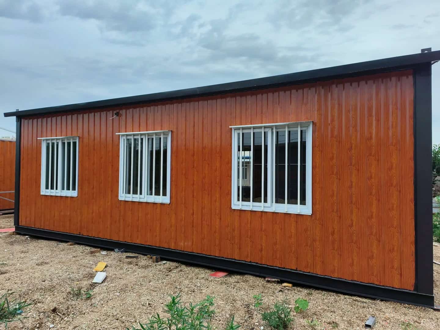 A Container Housing unit with a wooden - textured orange exterior and black framing. It features three white - framed windows and is situated on a gravel surface under a cloudy sky.