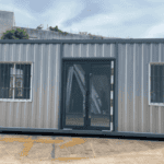 A compact grey container office with two small windows and a central glass door, placed outdoors on a concrete surface.