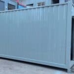 Light - gray container for refugee camp, with a door and window having metal bars, placed on a concrete surface.