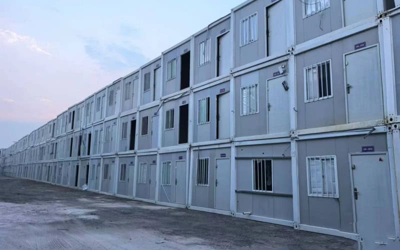 Rows of light - gray container refugee camp units with doors and windows, some open, arranged in a long line under a partly - cloudy sky.