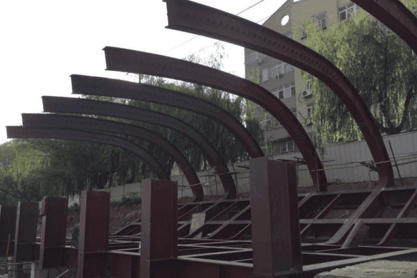 A partially built steel frame structure with curved beams, surrounded by trees and an urban backdrop.