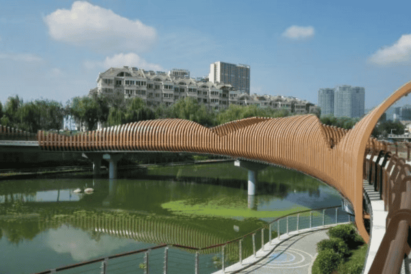 A wavy pedestrian bridge with a wooden aesthetic, crossing over a calm river with residential buildings in the background.