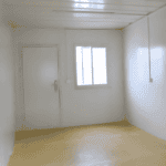 A simple, clean interior of a container unit featuring white walls, a single door, and a small window with ample natural light.