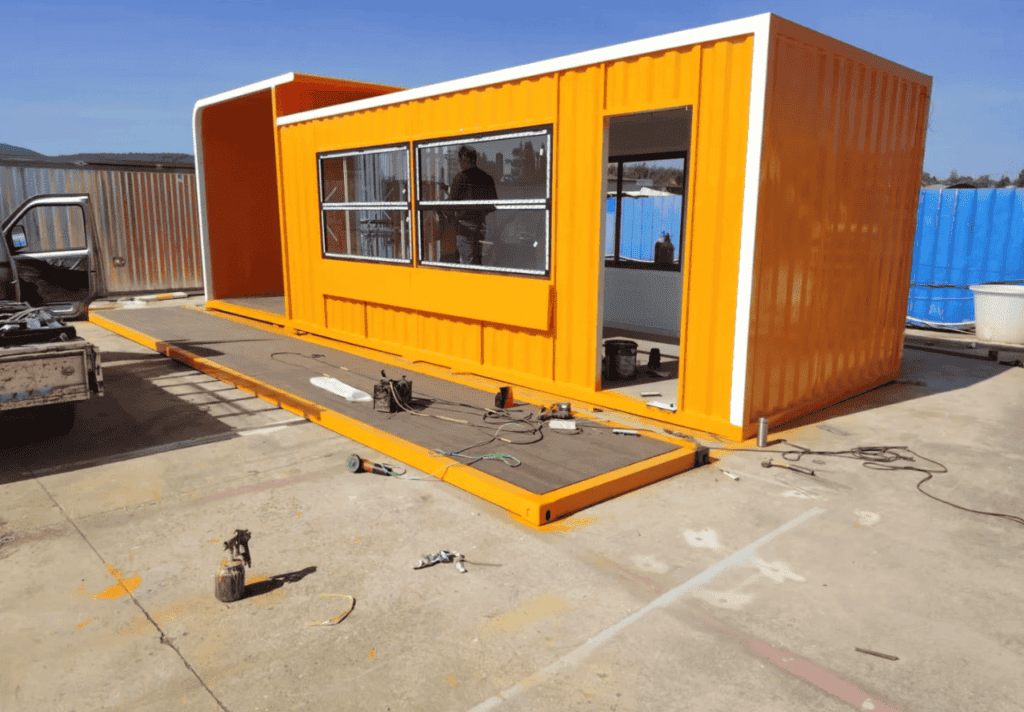 A bright orange detachable container house with large windows and an extended floor platform, under construction in an open area.