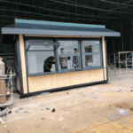 A small modular container unit with wood paneling and large windows, being assembled by workers in an industrial setting.