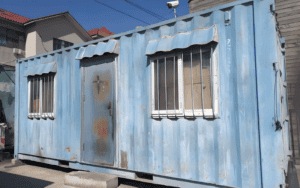 A blue container home with visible rust patches, situated outdoors near buildings.