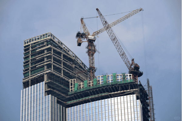 A skyscraper under construction, with cranes and scaffolding visible near the top against a cloudy sky.