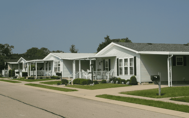 A row of prefab homes with pastel - colored exteriors, white porches, and neatly trimmed lawns along a quiet street. 