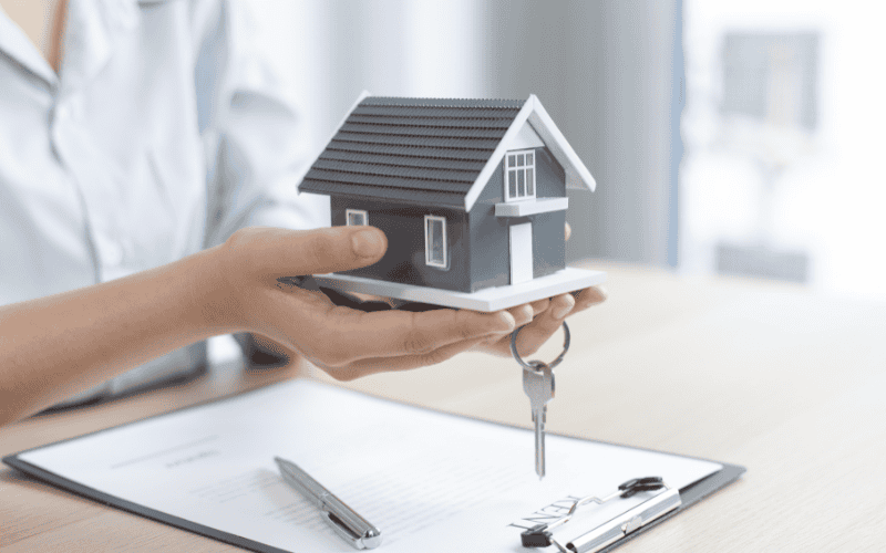 A person holds a small prefab home model over a clipboard with papers and a keychain, suggesting themes of housing and property.