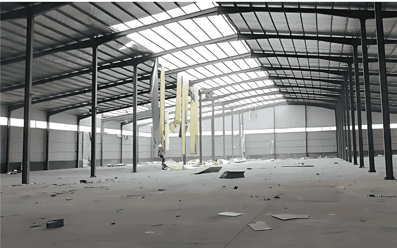 Wide, empty warehouse interior showing steel structure with a gabled roof, partially installed panels, and scattered construction debris.