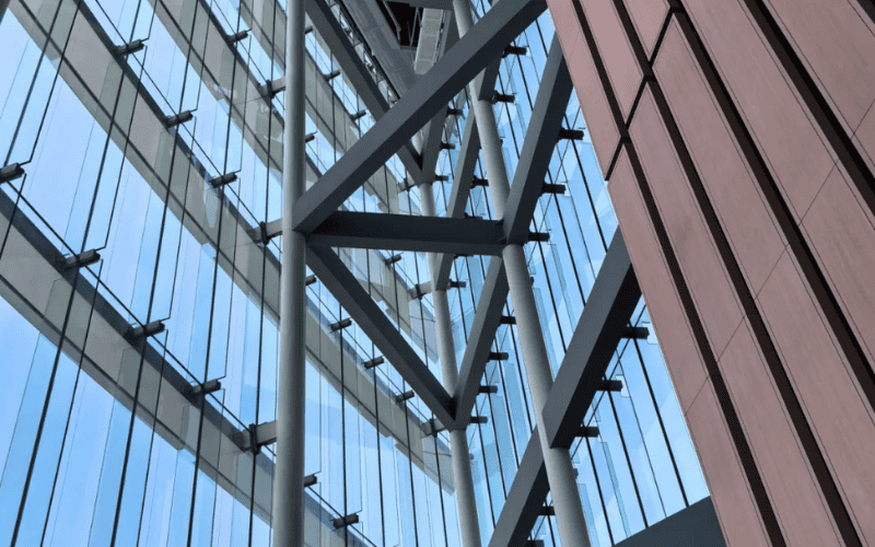 Close-up of a modern steel and glass structure, emphasizing angular beams and sleek architectural design, with a clear blue sky visible through the glass.