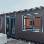 A dark brown expandable container house with large windows and a glass door, displayed outdoors near another modular unit.