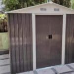 A single small farm equipment shed with a gable roof, ventilation openings, and double doors, placed near a fence.