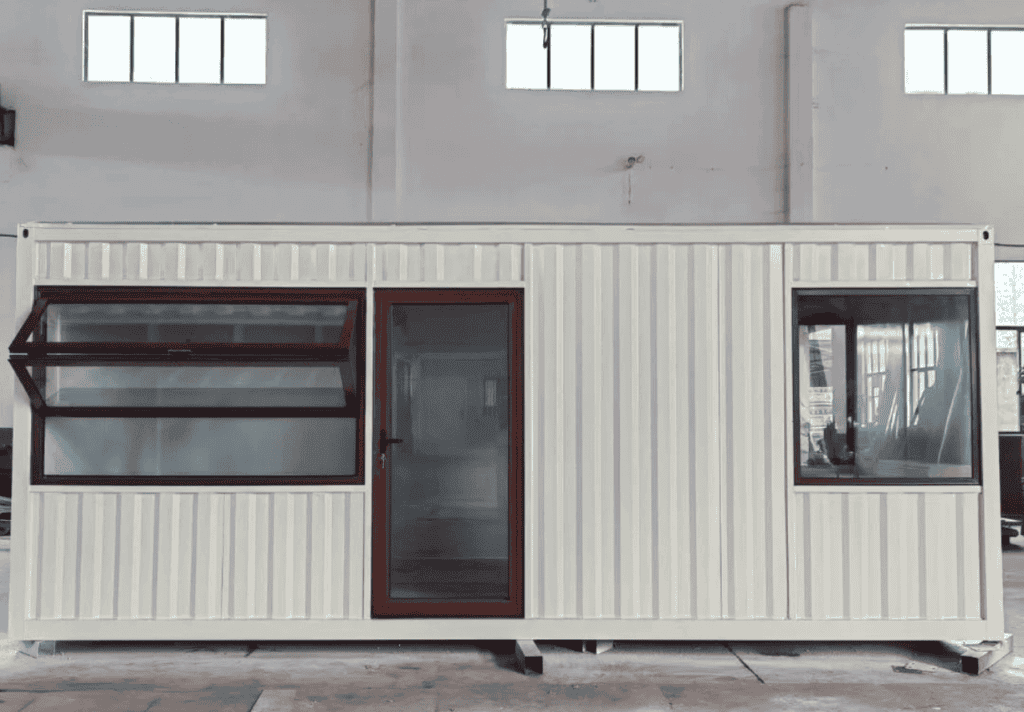 A white flat-pack container house with a clean design, featuring large windows and a glass door, showcased inside a spacious warehouse.