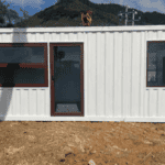 A white flat-pack container house placed outdoors, with a modern layout, large windows, and a scenic background.