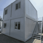 A two-story flat-pack container house with multiple windows and an exterior staircase, assembled on a concrete surface under bright sunlight.