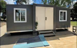 A dark gray foldable container house with white windows and a double front door, featuring a small wooden step and an outdoor rug for access.