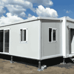 A white folding container house with a raised foundation, sliding doors, and windows, placed outdoors under a sunny sky.