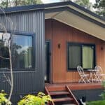 A dark - hued and wooden gable - roof portable cabin, having large windows, a wooden deck with white chairs and a table, and surrounded by greenery.