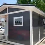 A gray gable - roof portable cabin with a front porch, showcasing a window with circular patterns and an open - style window on the side.