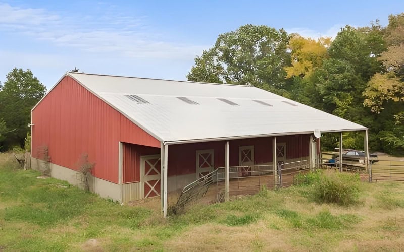 A red metal horse barn with a white roof and open - front stalls, surrounded by grass and trees under a blue sky.