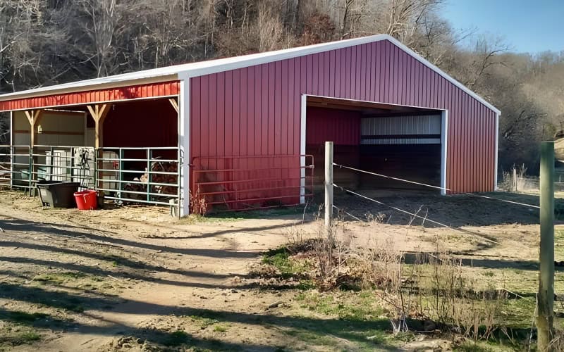 Another red metal horse barn with a white trim, one large opening and an attached shelter area, located on a dirt ground with some bushes nearby.