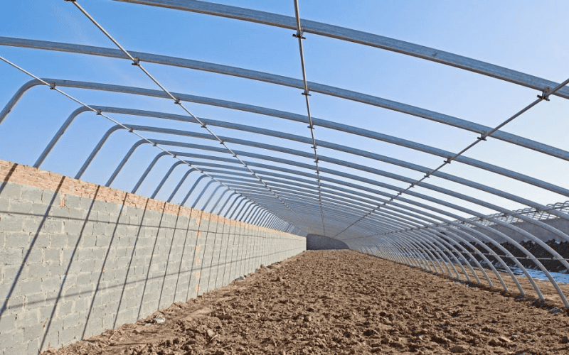 The framework of a pig house with arched metal beams and a brick - wall base, over a dirt - filled floor under a clear blue sky.