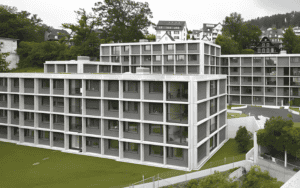 Multi - story temporary buildings with modern design, surrounded by greenery and other structures in the background.