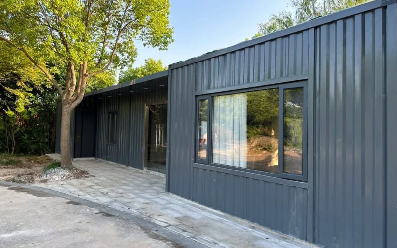 A dark grey shipping container office with large windows and a glass door, situated next to a tree on a paved area. The building is surrounded by greenery, giving it a modern and integrated look.