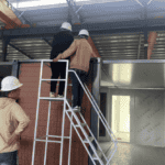 Workers inspecting a modular prefab house with a ladder placed against its side, highlighting construction details in a factory setting.