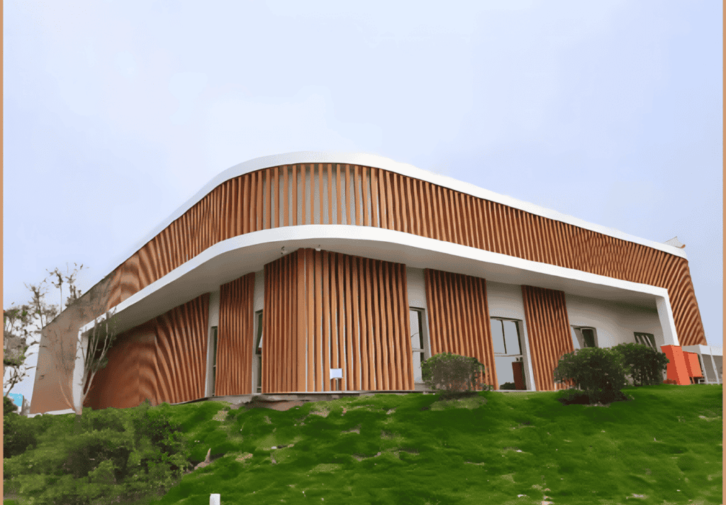 A modern light steel building with a wavy roofline and vertical wooden slats, surrounded by lush green landscaping.